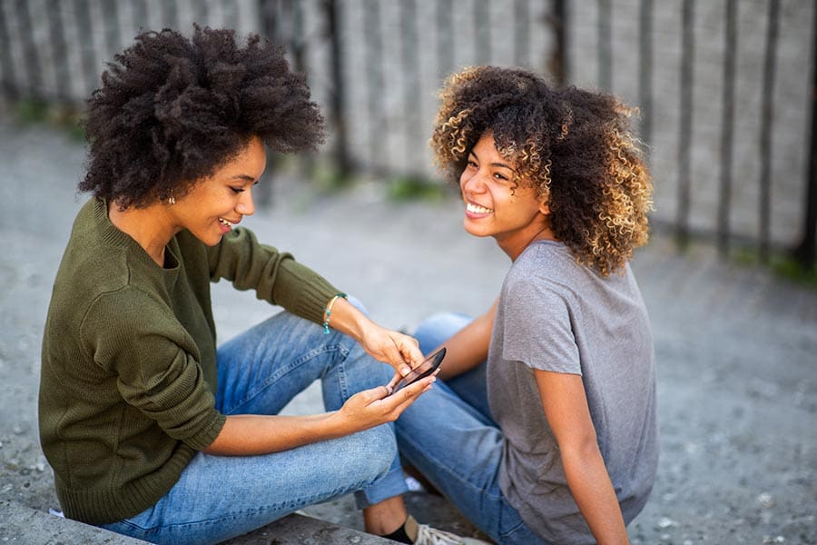 Two young girls sitting on stone steps, both smiling. One girl is using a phone while the other looks on, enjoying each other's company