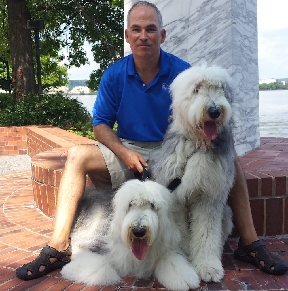 Jim O'Brien sitting with his two large dogs by the river, posing with the scenic river view in the background