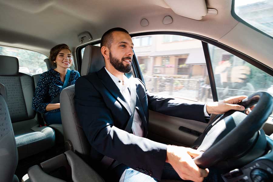 A man in a suit driving a car with a woman sitting in the back seat wearing a blue dress, both of them smiling